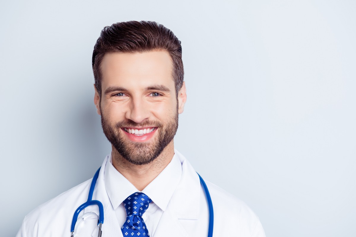 How a Geriatric Dentist Can Improve Your Care Professional male dentist in white coat with stethoscope smiling at camera against light blue background.