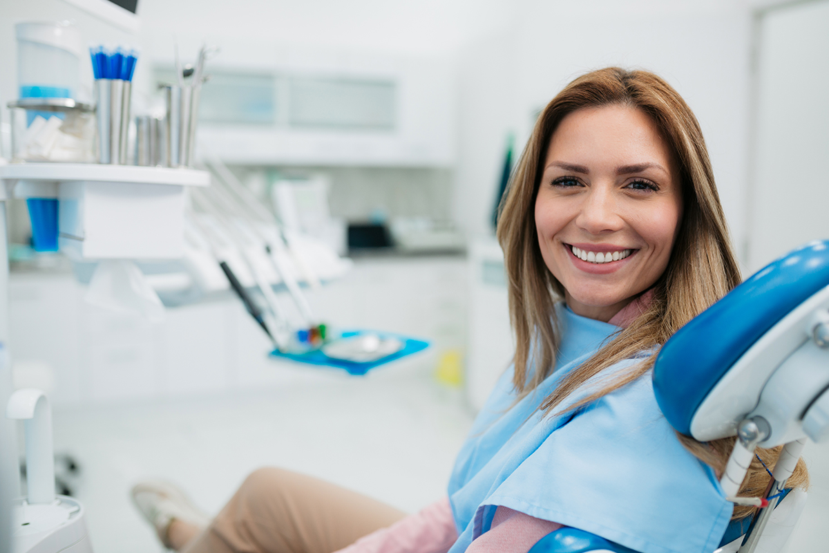 Female dentist smiling in modern dental office operatory with equipment visible in background.